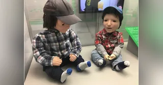 Dos robots Kaspar sentados, fotografiados como parte de una exposición.