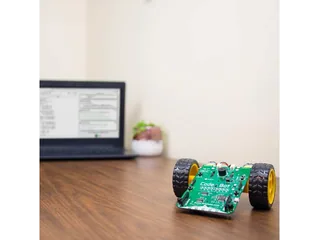 CodeBot con placa electrónica verde y dos ruedas sobre una mesa junto a un ordenador.