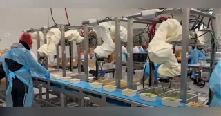 Robots de Chef Robotics con fundas protectoras trabajando junto a personas en una línea de preparación de bandejas de comida.