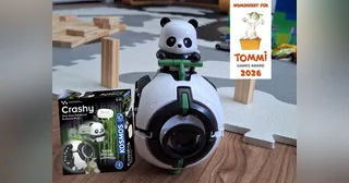 Fotografía promocional oficial de Crashy completo sobre el suelo, junto a su caja y el anuncio de su nominación al TOMMI 2026.