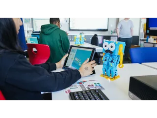 Fotografía oficial de Marty V2 azul y amarillo sobre una mesa de aula, junto a una persona que utiliza su interfaz de programación en una tableta.