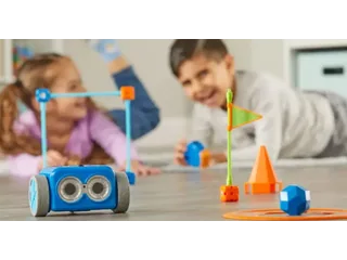 Botley 2.0 azul sobre un suelo de madera, con piezas de un circuito y niños jugando al fondo.