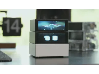 Fotograma del vídeo oficial de Cubie completo, con pantallas expresivas y base blanca, sobre un escritorio.