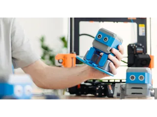 Una persona sostiene un HP Otto azul y gris con patas y ruedas de la configuración Walk & Roll, delante de una impresora 3D, en una fotografía de Moravia.