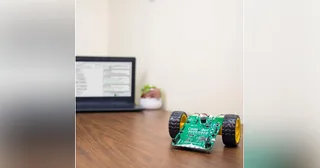 CodeBot con placa electrónica verde y dos ruedas sobre una mesa junto a un ordenador.