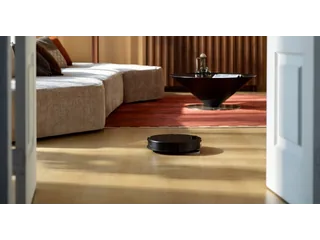 Fotografía promocional oficial de un Roborock Saros 20 Neo negro sobre suelo de madera, visto a través de una puerta frente a un sofá y una alfombra.