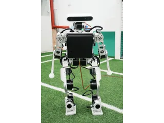 Fotografía oficial de MOS9.2 completo, de frente, con estructura metálica, brazos y pies sobre un campo de fútbol robótico.