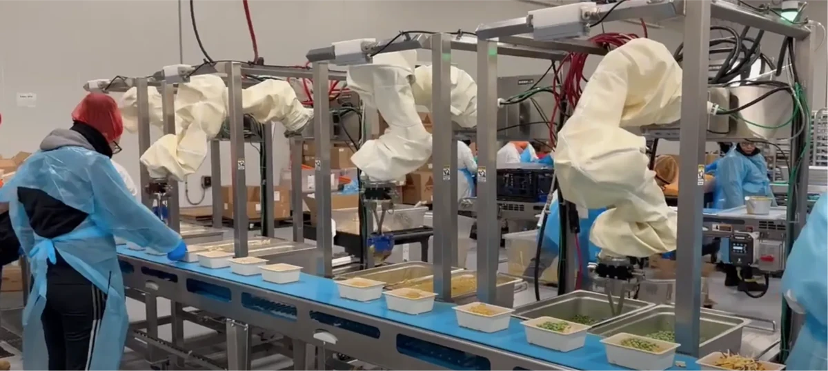 Robots de Chef Robotics con fundas protectoras trabajando junto a personas en una línea de preparación de bandejas de comida.