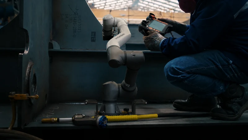 Robot de soldadura ArcLift GO de HD Hyundai trabajando junto a un operario dentro de una estructura de acero en un astillero.